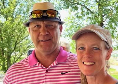 A couple poses outdoors, surrounded by greenery, wearing stylish hats and casual attire. Sunny day, cheerful atmosphere